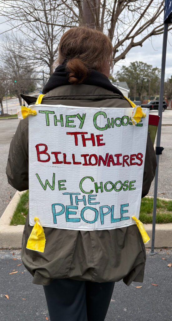 Back of woman with sign that says "They Choose the Billionaires. We Choose the People"
